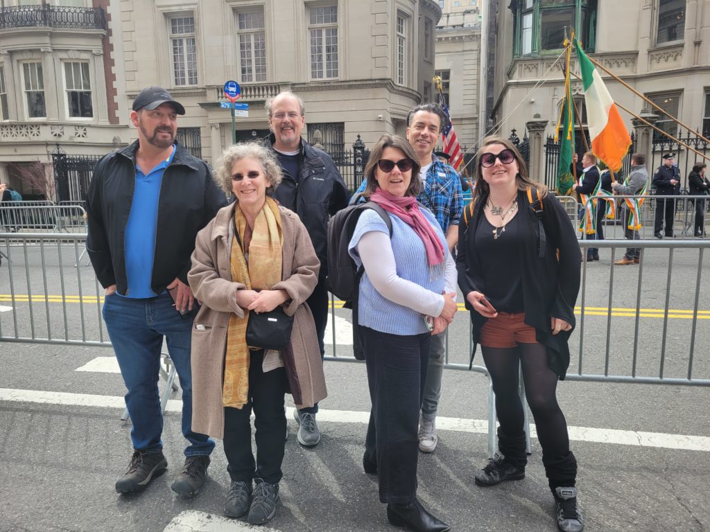 Three men and three women standing in front of an Irish flag at a St Patrick's Day parade.
